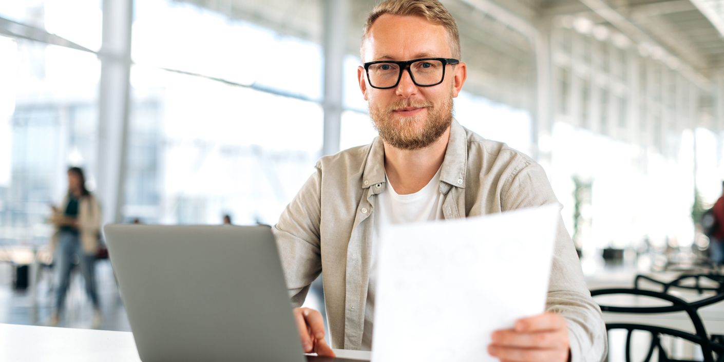 Freundlicher motivierter erfolgreicher kaukasischer Mann, mit Brille, IT-Spezialist, Firmen-Seo, Manager, Freiberufler, sitzt an einem Schreibtisch mit einem Laptop in Coworking, arbeitet mit Dokumenten, schaut in die Kamera, lächelt